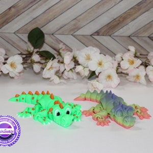 May include: Two toy alligators in vibrant colours are shown. One is bright green with orange spikes, the other a gradient of pink, green, and blue. Both have cartoon eyes and are lying down on a white surface.