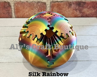 Op de afbeelding: Een veelkleurige, iriserende, 3D-geprinte bol met een tandwielachtig ontwerp. De bol rust op een houten oppervlak. De tekst "Aly's Craft Boutique" is zichtbaar in de afbeelding. De tekst "Silk Rainbow (Color Transitions will vary between items ordered)" is ook zichtbaar in de afbeelding.