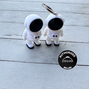 Può includere: Due portachiavi a forma di astronauta bianchi con visiere e dettagli neri. Un portachiavi ha un anello argentato attaccato. I portachiavi sono su una superficie di legno grigio chiaro. Un logo Venditore Autorizzato in bianco e nero è nell'angolo in basso a destra.