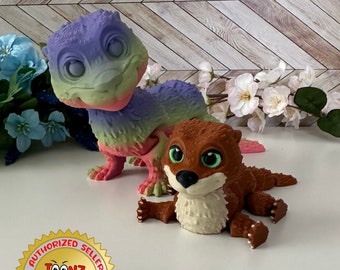 May include: Two 3D-printed figurines: a colorful lizard-like creature and a brown otter with green eyes. The lizard is primarily purple, green, and pink. The otter has a white chest and is lying down. The Toonz Factory logo is visible.