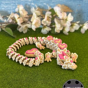 Puede incluir: Una figurita de dragón hecha a mano de arcilla polimérica con una base blanca y marrón y detalles de corazón rosa. El dragón está enroscado en una posición de dormir sobre una superficie verde.