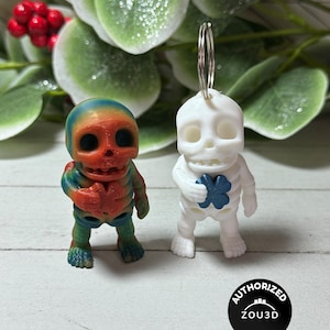 Op de afbeelding: Twee kleine skeletbeeldjes. Eén is wit met een blauwe klaver met vier bladeren, de andere is veelkleurig met rode, blauwe en groene tinten. Beide hebben sleutelhangers. De achtergrond omvat groene bladeren en een wit houten oppervlak.