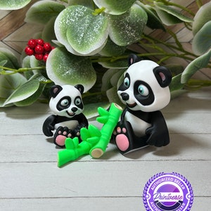 Peut inclure: Deux figurines de pandas, une grande et une petite, avec une fourrure noire et blanche et des accents roses sur les pieds. Ils sont assis près de tiges de bambou vertes. Le fond comprend des feuilles vertes et des baies rouges.