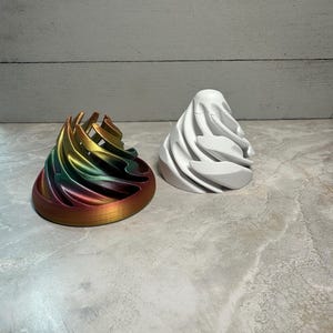 May include: Two 3D printed objects, one is a white cone-shaped object with a swirling design, the other is a multi-coloured object with a swirling design in shades of green, gold, and red.