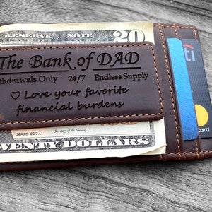 May include: A brown leather wallet with a money clip and a card holder. The wallet has the text "The Bank of DAD" engraved on the front. The text also says "Withdrawals Only 24/7 Endless Supply Love your favorite financial burdens".