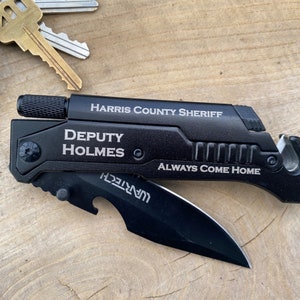 May include: Black multi-tool with a silver blade and a black handle. The handle has the text "HARRIS COUNTY SHERIFF" and "DEPUTY HOLMES" engraved on it. The tool also has a bottle opener and a seatbelt cutter. The text "ALWAYS COME HOME" is engraved on the handle.
