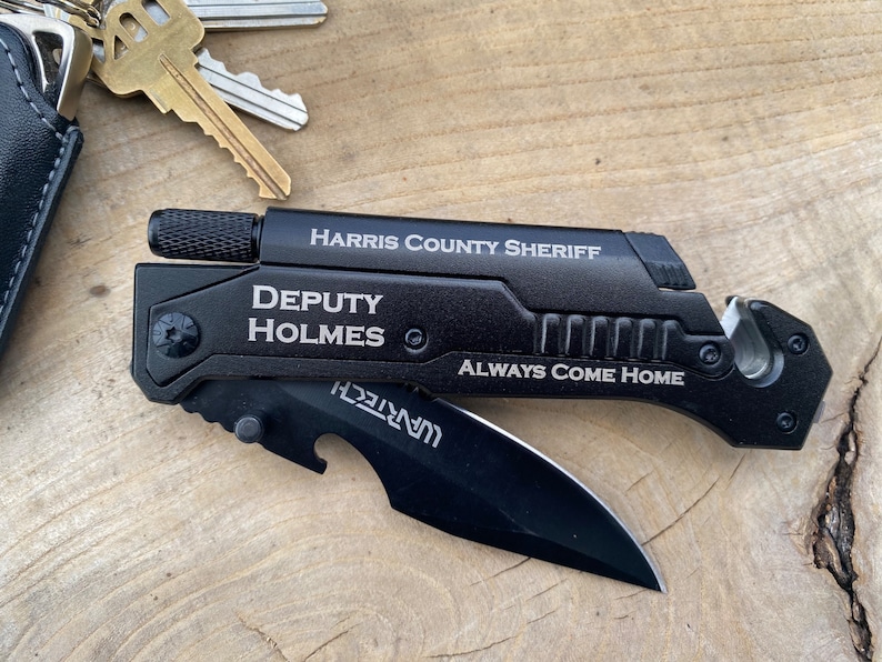 May include: Black multi-tool with a silver blade and a black handle. The handle has the text "HARRIS COUNTY SHERIFF" and "DEPUTY HOLMES" engraved on it. The tool also has a bottle opener and a seatbelt cutter. The text "ALWAYS COME HOME" is engraved on the handle.
