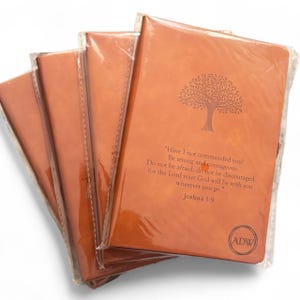 May include: Several brown leather journals are stacked, each wrapped in clear plastic. The front journal features a tree design and text from Joshua 1:9. The journals have a smooth texture and are a warm, earthy tone.