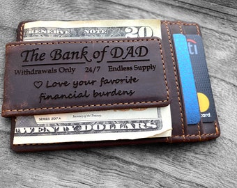 May include: A dark brown leather money clip wallet with orange stitching. The front is engraved with "The Bank of DAD", "Withdrawals Only 24/7 Endless Supply", and "Love your favorite financial burdens" with a heart icon. It holds a twenty dollar bill.