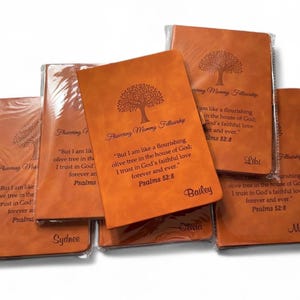 May include: A collection of orange-brown journals with embossed tree designs and text. Each journal is wrapped in clear plastic. The text includes the phrase "Flourishing Mummy Fellowship" and a Bible verse.