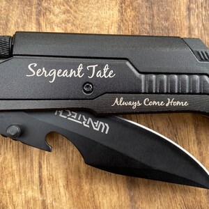 May include: Black multi-tool knife with a black blade and a bottle opener. The knife has a safety feature and a belt cutter. The knife is engraved with the text "Sergeant Tate Always Come Home".