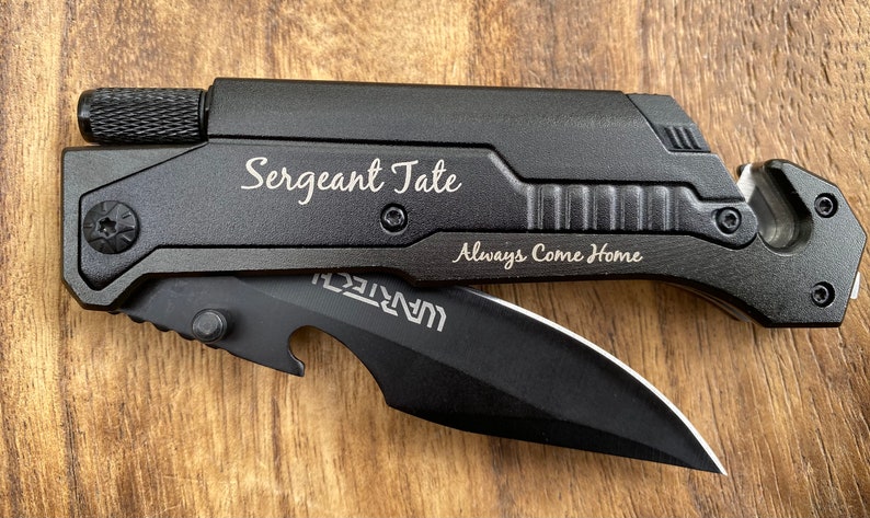 May include: Black multi-tool knife with a black blade and a bottle opener. The knife has a safety feature and a belt cutter. The knife is engraved with the text "Sergeant Tate Always Come Home".