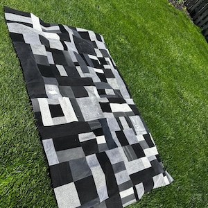 May include: A rectangular quilt with a patchwork design in shades of black, grey, and white. The quilt is made of various fabric squares and rectangles, creating a geometric pattern. The quilt is lying on a green lawn.