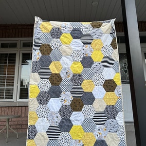May include: A yellow, grey, and white quilt with a honeycomb pattern. The quilt is made of different fabrics with various patterns, including stripes, gingham, and floral prints.
