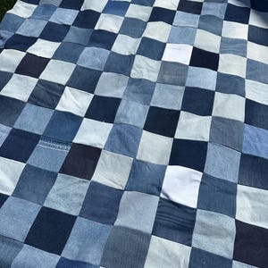 May include: A patchwork quilt made from squares of denim in various shades of blue and white. The quilt has a checkerboard pattern, with each square approximately 20 cm by 20 cm. The quilt is lying on a grassy surface.