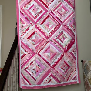 May include: A pink and white quilt with a diamond pattern. The quilt is made of various fabrics with different patterns and textures.