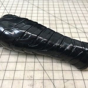 May include: Black plastic forearm armor with a textured, geometric design. The armor is curved to fit the shape of the arm.