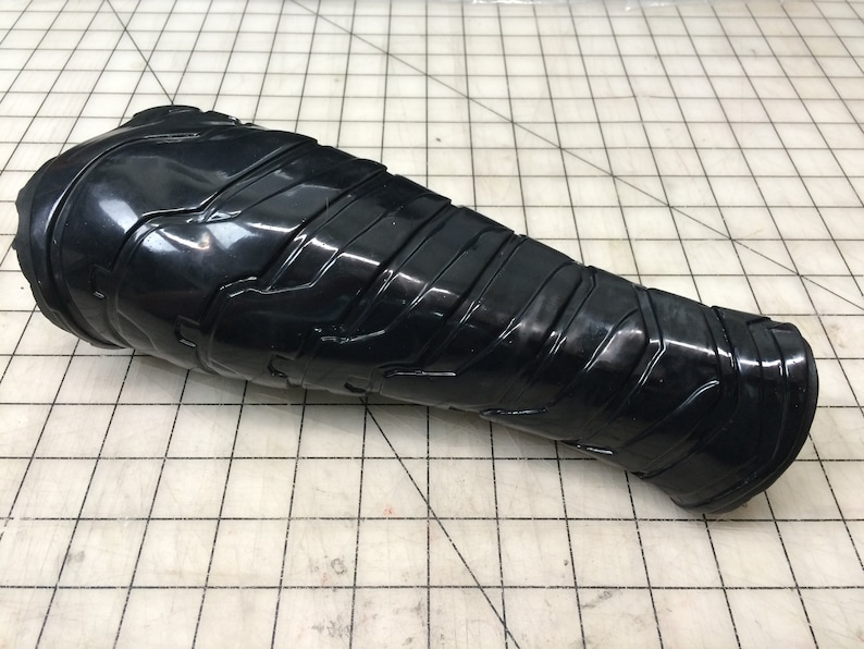 May include: Black plastic forearm armor with a textured, geometric design. The armor is curved to fit the shape of the arm.