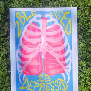 May include: A poster with a blue background features a pink illustration of lungs and rib cage. The words "Blessed 2B Depressed" are written in yellow, arching around the top and bottom of the illustration.