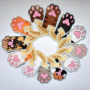 Peut inclure: Un ensemble de 12 autocollants en forme de patte de chat avec un fond bleu clair et une bannière dorée qui dit "Toebean Farm". Les empreintes de pattes sont de différentes couleurs et motifs, notamment orange, noir, marron et gris.