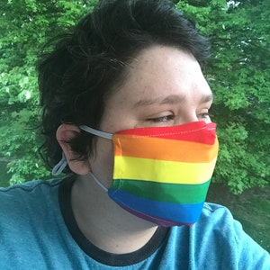 May include: A face mask with a rainbow stripe design and white elastic ear loops. The mask displays horizontal bands of red, orange, yellow, green, blue, and purple. The person is wearing a blue t-shirt.