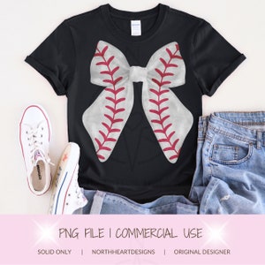 May include: A black t-shirt with a white and red baseball-themed bow on the front. The bow is made to look like a baseball with red stitching. The shirt is laying on a white surface with a pair of white sneakers and blue jeans.
