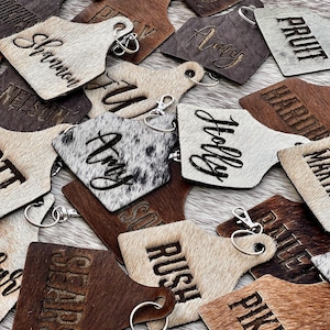 Puede incluir: Una colección de llaveros personalizados con varios nombres y palabras grabadas. Los llaveros son de madera y piel de vaca, con llaveros plateados. Los colores varían del marrón claro al marrón oscuro.