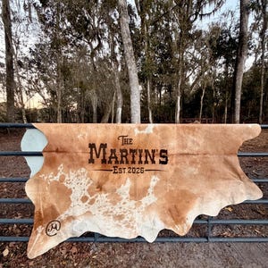 May include: A cowhide rug with a brown and white spotted pattern. The rug is draped over a metal fence. The text "The Martin's Est. 2026" is printed in black on the rug.