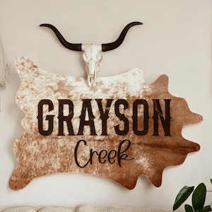 Może przedstawiać: Rustykalna dekoracja ścienna w kształcie skóry krowy z napisem "GRAYSON Creek" w ciemnobrązowych literach. Nad znakiem znajduje się czaszka krowy z długimi rogami. Znak jest zamontowany na ścianie w neutralnym kolorze.