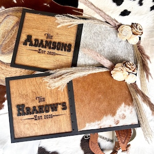 Op de afbeelding: Twee houten familienaamborden met zwarte frames. Eén bord toont de naam "The Adamsons" en "Est. 2025", de andere "The Krakow's" en "Est. 2025". De borden zijn versierd met gedroogde bloemen en pampasgras.