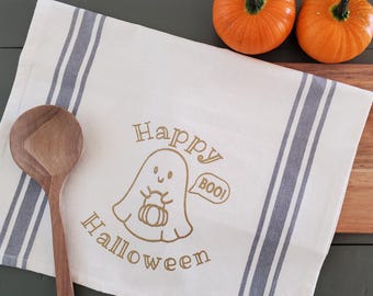 Puede incluir: Un paño de cocina crema con temática de Halloween con rayas azules y un gráfico de fantasma que dice "Happy Halloween" y "Boo!". Dos calabazas naranjas y una cuchara y tabla de cortar de madera también están en la imagen.