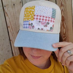 May include: A baseball cap with a cream-colored crown and a light blue denim brim. The front panel features a rectangular patchwork design composed of various fabric squares in yellow, red gingham, blue gingham, and floral patterns.