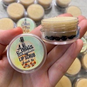 May include: Clear plastic lip scrub containers with a light brown scrub and dark brown beads. The label reads "Artisan Bath Co. LIP SCRUB Bubble Tea 0.5 oz - 14 g". Other containers are visible in the background.