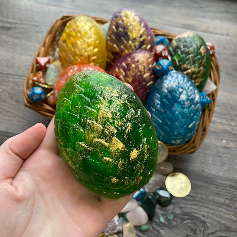 Peut inclure: Une collection de savons en forme d'&oelig;uf de dragon vert, bleu, violet et jaune dans un panier en osier. Les savons sont d&eacute;cor&eacute;s de paillettes dor&eacute;es.