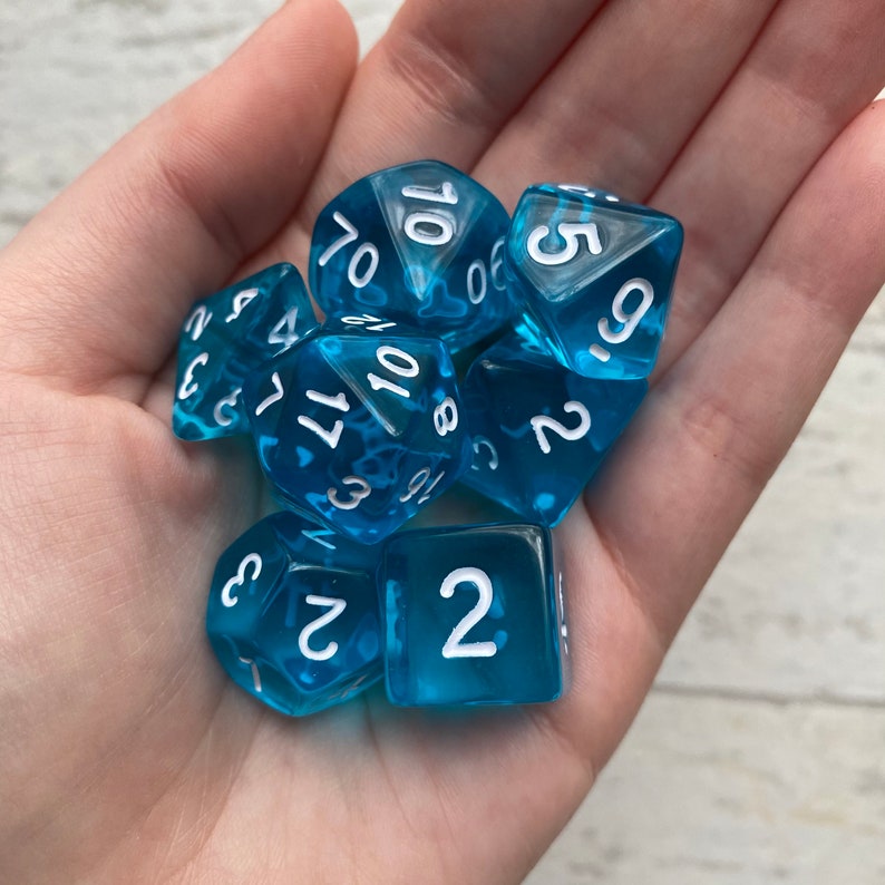 Peut inclure: Un ensemble de six d&eacute;s bleus avec des chiffres blancs. Les d&eacute;s sont dans une main, et les chiffres visibles sont 3, 4, 11, 10, 2 et 5.