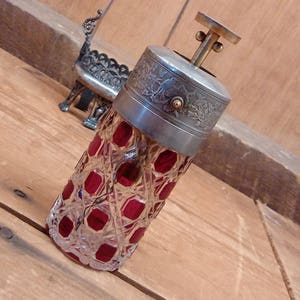 May include: A silver and glass bottle stopper with a red glass body and a silver top with an ornate design. The stopper is on a wooden surface.