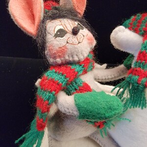 Puede incluir: Una figura de rat&oacute;n de fieltro hecha a mano con grandes orejas rosas, que lleva una bufanda a rayas rojas y verdes y mitones verdes. El rat&oacute;n tiene pelo negro y un sombrero rojo. Una decoraci&oacute;n festiva navide&ntilde;a.