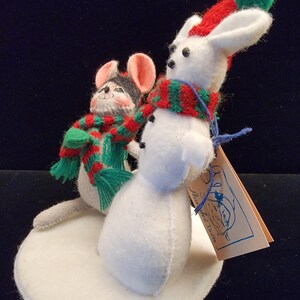 Puede incluir: Decoraci&oacute;n navide&ntilde;a de fieltro con un mu&ntilde;eco de nieve blanco y un rat&oacute;n, ambos con bufandas festivas. El mu&ntilde;eco de nieve tiene un gorro rojo con un pomp&oacute;n verde. El rat&oacute;n tiene una cabeza gris y negra y orejas rosas. Est&aacute;n sobre una base de fieltro blanco.