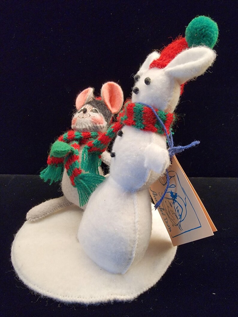 Puede incluir: Decoraci&oacute;n navide&ntilde;a de fieltro con un mu&ntilde;eco de nieve blanco y un rat&oacute;n, ambos con bufandas festivas. El mu&ntilde;eco de nieve tiene un gorro rojo con un pomp&oacute;n verde. El rat&oacute;n tiene una cabeza gris y negra y orejas rosas. Est&aacute;n sobre una base de fieltro blanco.