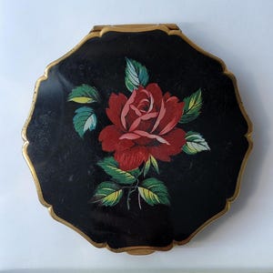 May include: A vintage black compact with a gold trim and a red rose painted on the lid. The rose has green leaves and a detailed design. The compact has a unique, scalloped shape.
