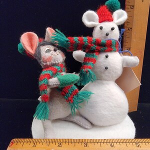 Puede incluir: Un adorno navide&ntilde;o caprichoso con dos ratones de fieltro y un mu&ntilde;eco de nieve. Los ratones llevan bufandas y gorros a rayas rojas y verdes. El mu&ntilde;eco de nieve es blanco con ojos de bot&oacute;n negros. El adorno est&aacute; sobre una base blanca.