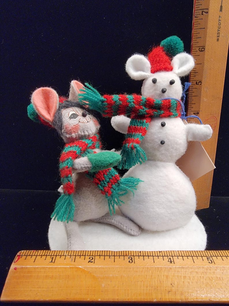 Puede incluir: Un adorno navide&ntilde;o caprichoso con dos ratones de fieltro y un mu&ntilde;eco de nieve. Los ratones llevan bufandas y gorros a rayas rojas y verdes. El mu&ntilde;eco de nieve es blanco con ojos de bot&oacute;n negros. El adorno est&aacute; sobre una base blanca.