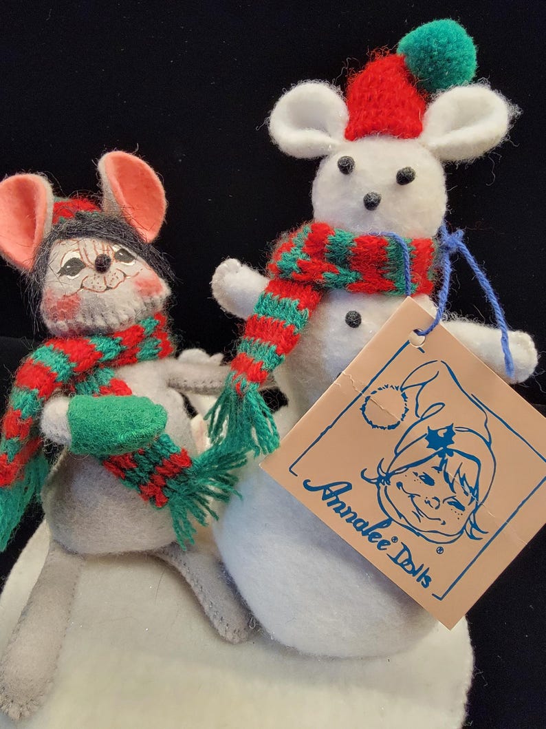 Puede incluir: Dos mu&ntilde;ecos de rat&oacute;n de peluche, uno gris y otro blanco, con bufandas y sombreros rojos y verdes. El rat&oacute;n blanco tiene un sombrero rojo con un pomp&oacute;n verde. El rat&oacute;n gris tiene un sombrero negro. Una etiqueta dice "Annalee Dolls".