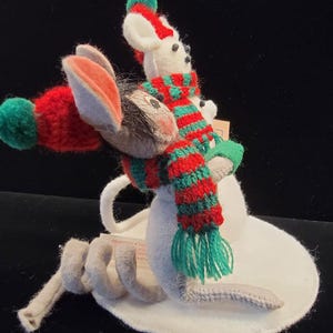 Puede incluir: Un adorno navide&ntilde;o caprichoso con un rat&oacute;n y un mu&ntilde;eco de nieve. El rat&oacute;n lleva un gorro rojo con un pomp&oacute;n verde y una cola gris. El mu&ntilde;eco de nieve lleva una bufanda a rayas rojas y verdes. El adorno est&aacute; sobre una base blanca.