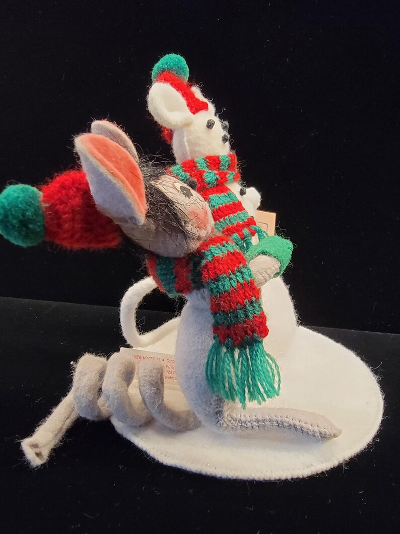 Puede incluir: Un adorno navide&ntilde;o caprichoso con un rat&oacute;n y un mu&ntilde;eco de nieve. El rat&oacute;n lleva un gorro rojo con un pomp&oacute;n verde y una cola gris. El mu&ntilde;eco de nieve lleva una bufanda a rayas rojas y verdes. El adorno est&aacute; sobre una base blanca.