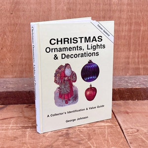 May include: A yellow book titled "Christmas Ornaments, Lights & Decorations" with a picture of Santa Claus, a purple glass ornament, and a red apple ornament. The book is a collector's identification and value guide by George Johnson.