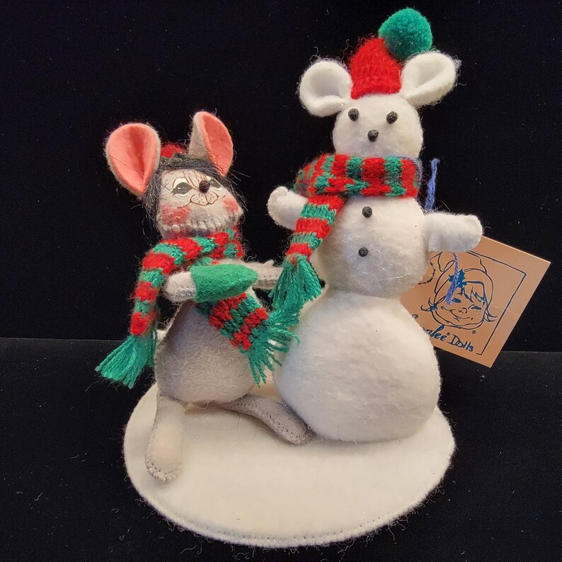 Puede incluir: Una decoraci&oacute;n navide&ntilde;a de fieltro con dos ratones. Un rat&oacute;n es gris con una bufanda y un gorro rojos y verdes, el otro es blanco con una bufanda y un gorro rojos y verdes, encima de un mu&ntilde;eco de nieve. La base es un c&iacute;rculo de fieltro blanco.