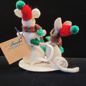 Puede incluir: Dos figuras de ratones de fieltro blanco con bufandas y gorros a rayas rojas y verdes con pompones verdes. Los ratones est&aacute;n sobre una base circular blanca. Una etiqueta dice "Made only by Annalee".