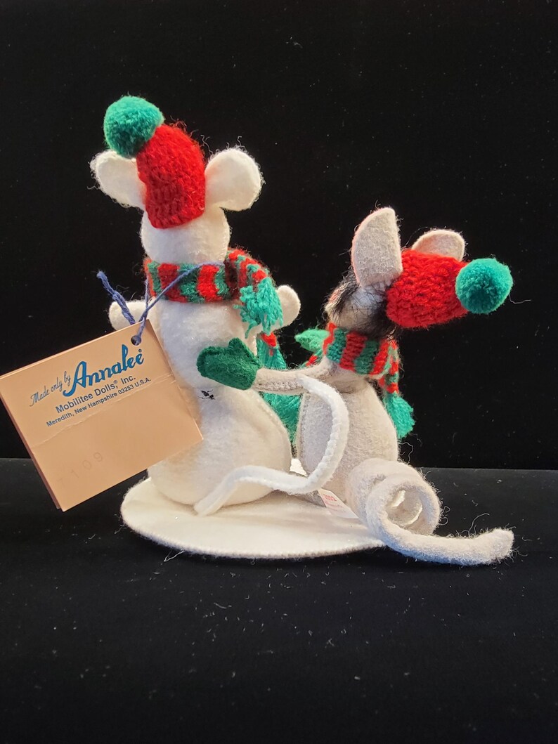 Puede incluir: Dos figuras de ratones de fieltro blanco con bufandas y gorros a rayas rojas y verdes con pompones verdes. Los ratones est&aacute;n sobre una base circular blanca. Una etiqueta dice "Made only by Annalee".
