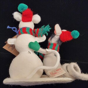 Puede incluir: Una caprichosa figura de mu&ntilde;eco de nieve con una bufanda a rayas rojas y verdes, un gorro a juego y guantes verdes. El mu&ntilde;eco de nieve est&aacute; hecho de fieltro blanco y tiene una larga cola rizada. Una etiqueta dice "2002" y "Amalie".